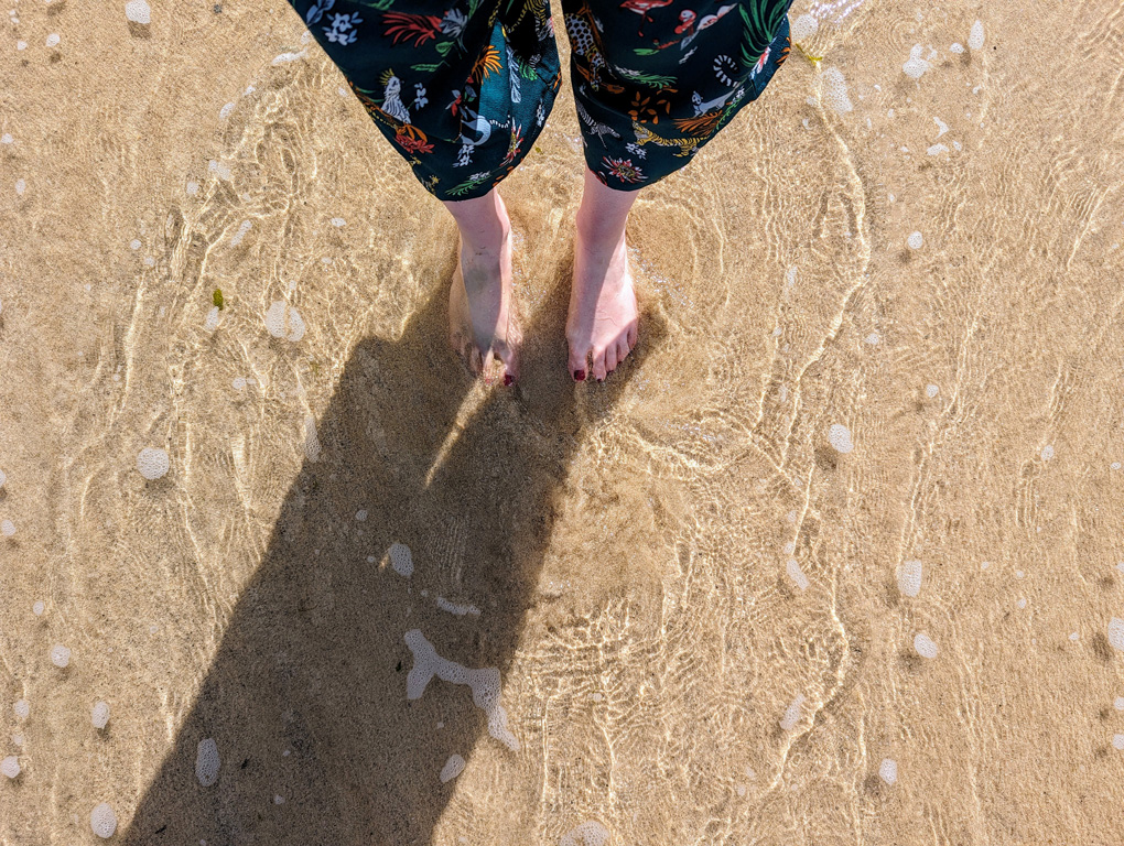 A view down to my feet partially buried in sand and with bright clear waves washing over them