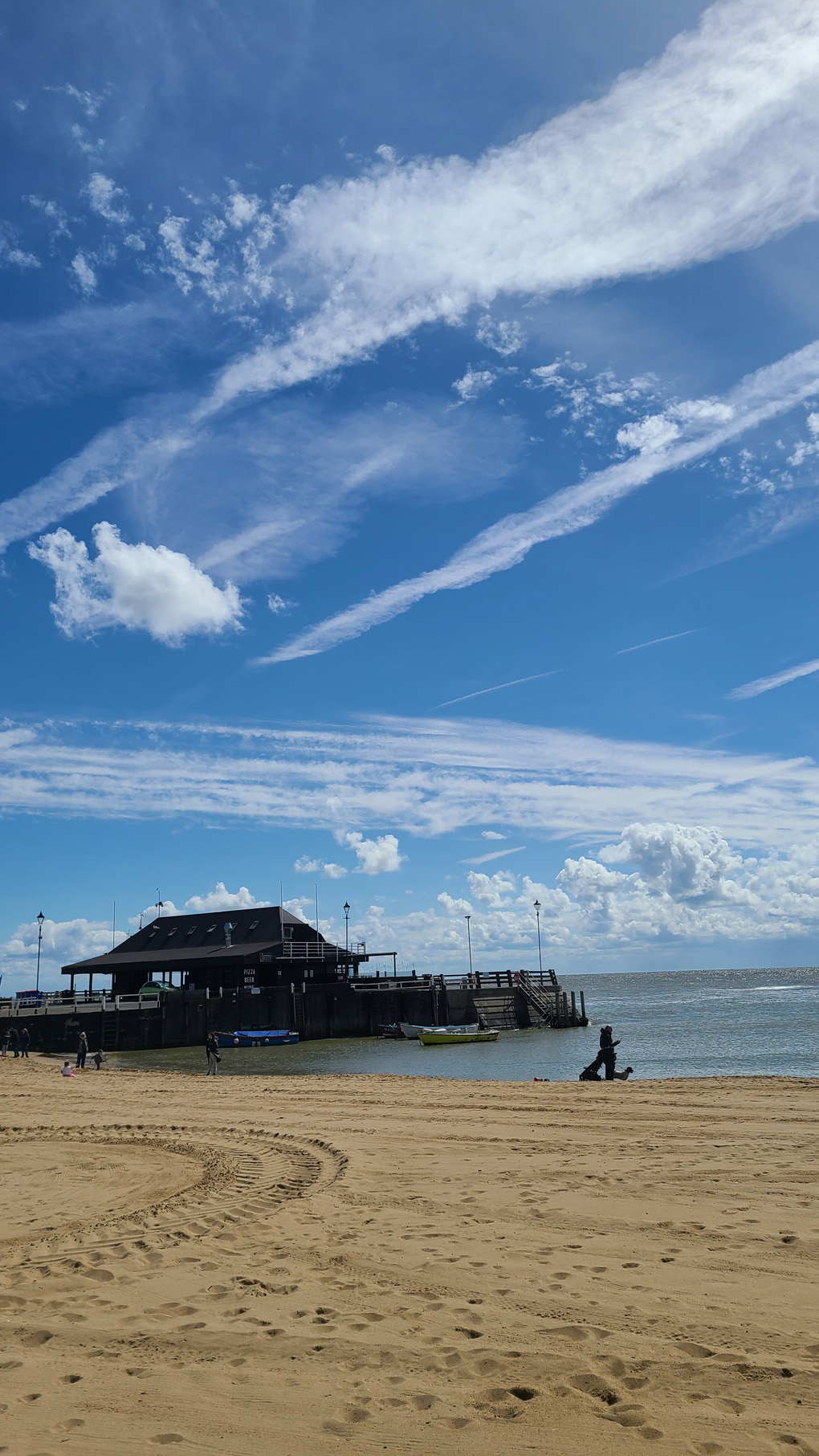 A bright Spring day. Lots of different white clouds against a blue sky, over a the sea and beach. There is a jetty with a large building on it.
