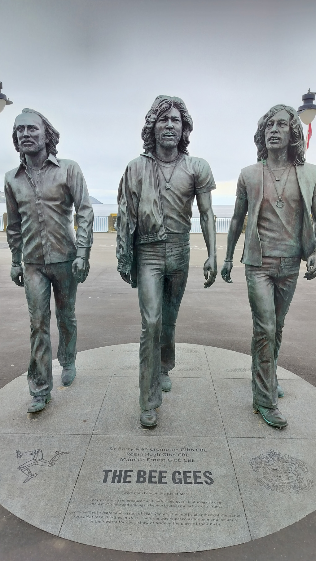 A statue of the three Bee Gees, walking in a line, on the promenade in Douglas. They are walking away from the sea, which is visible in the background.