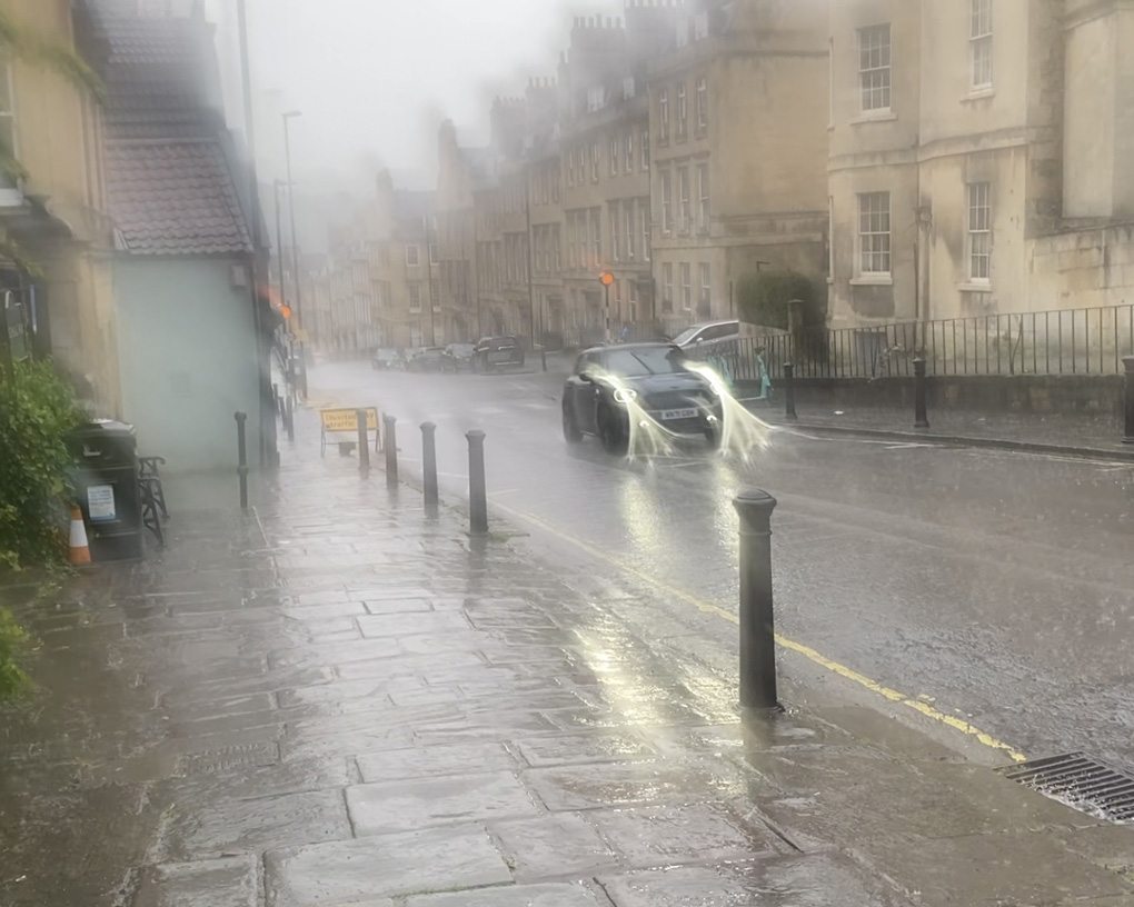 A view of a car driving up a street in Bath during torrential rain. Water has smeared the lens, leaving the picture blurry.