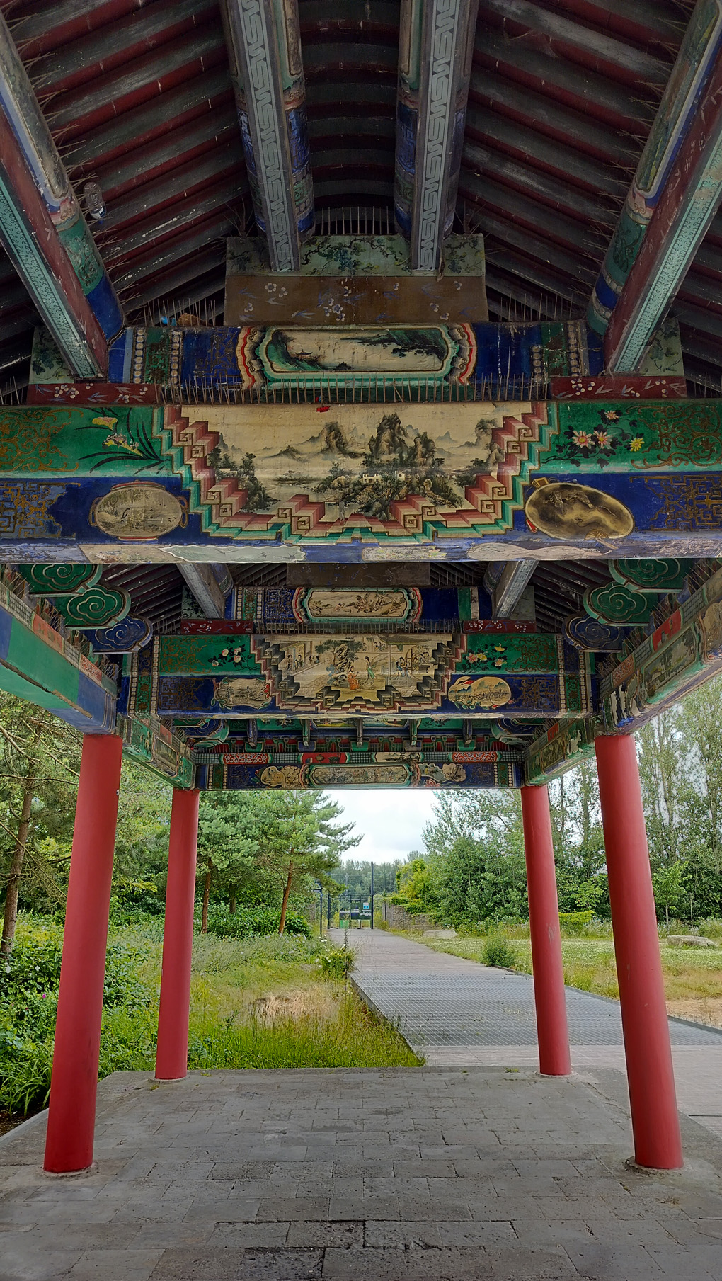 Standing inside a japanese-style pagoda with red pillars we see the roof, which has paintings onthe beams of mountains, lakes, flowers and people on it. Heading off into the distance is a path with overgrown grass on each side and a security gate at the end, which is closed.