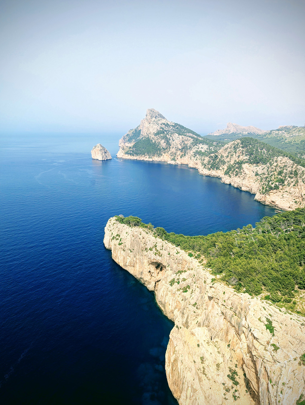 A high view of a winding coastline and roads following it's edge. The sky is a deep vivid blue and there is a summer hazy heat on the horizon