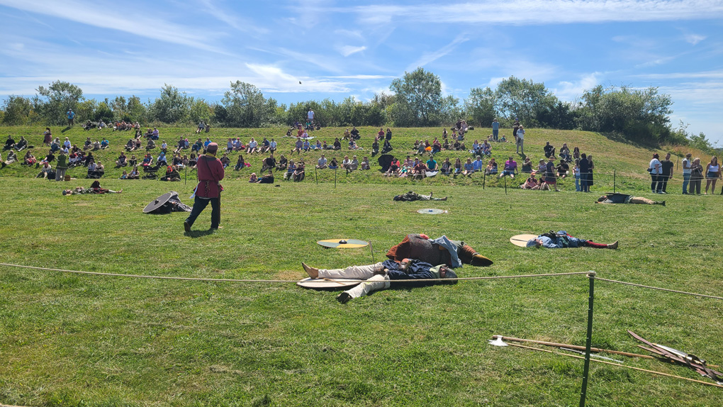 A grass amphitheater with a rope cordon, blue sky above. Spectators sit watching a Viking battle, many lay 