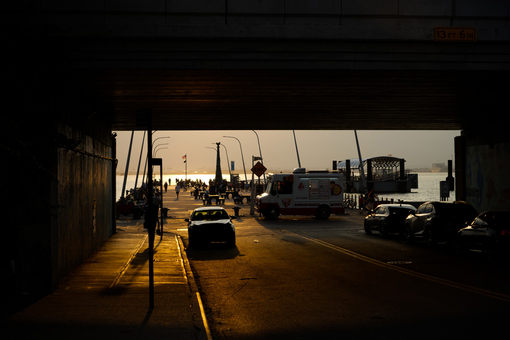 The Bay Ridge Pier in Brooklyn can be seen through a freeway underpass. Late afternoon light is gold and yellow tinted, and is reflecting sharply off an old sedan, and the white road markings. People on bikes and fishing can be seen on the pier itself in the background.