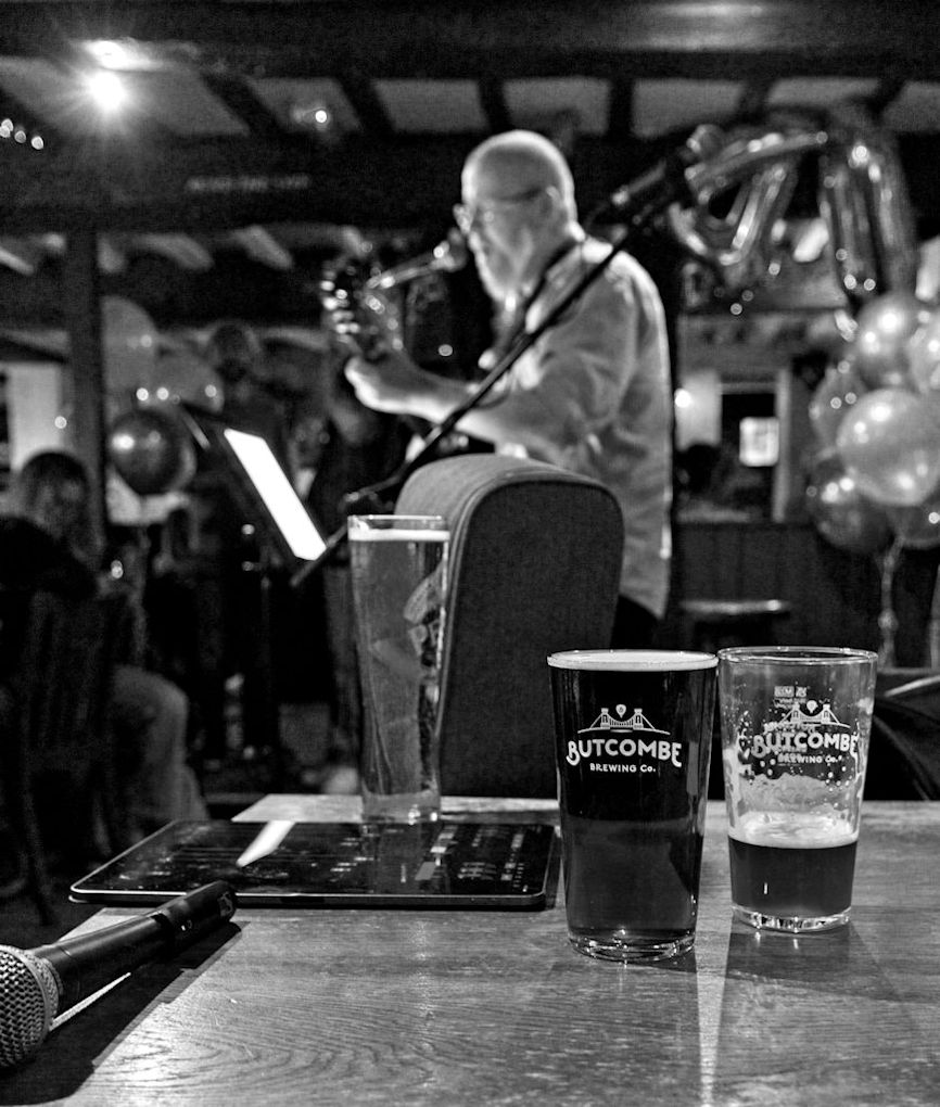 Black and White image with a single singer/guitarist at the mic in the middle ground but a shallow depth of field throws him out of focus with on the beer on the table at the front of the image being the only sharp part of the frame.