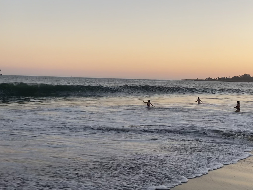 Large waves rolling in towards a beach at sunset. Three people are wading into the water towards the waves, arms outstretched