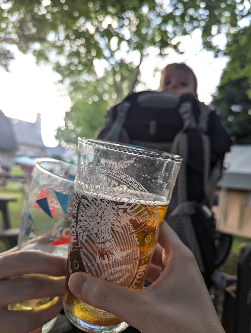 Two people holding half-drunk glasses of beer. On the background, our of focus, is a baby peering over the top of a backpack carrier