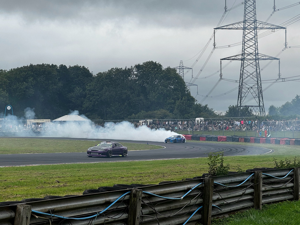 Two sports cars - purple in the foreground, blue in the background - drifting around a wide curve of a race track in rural England