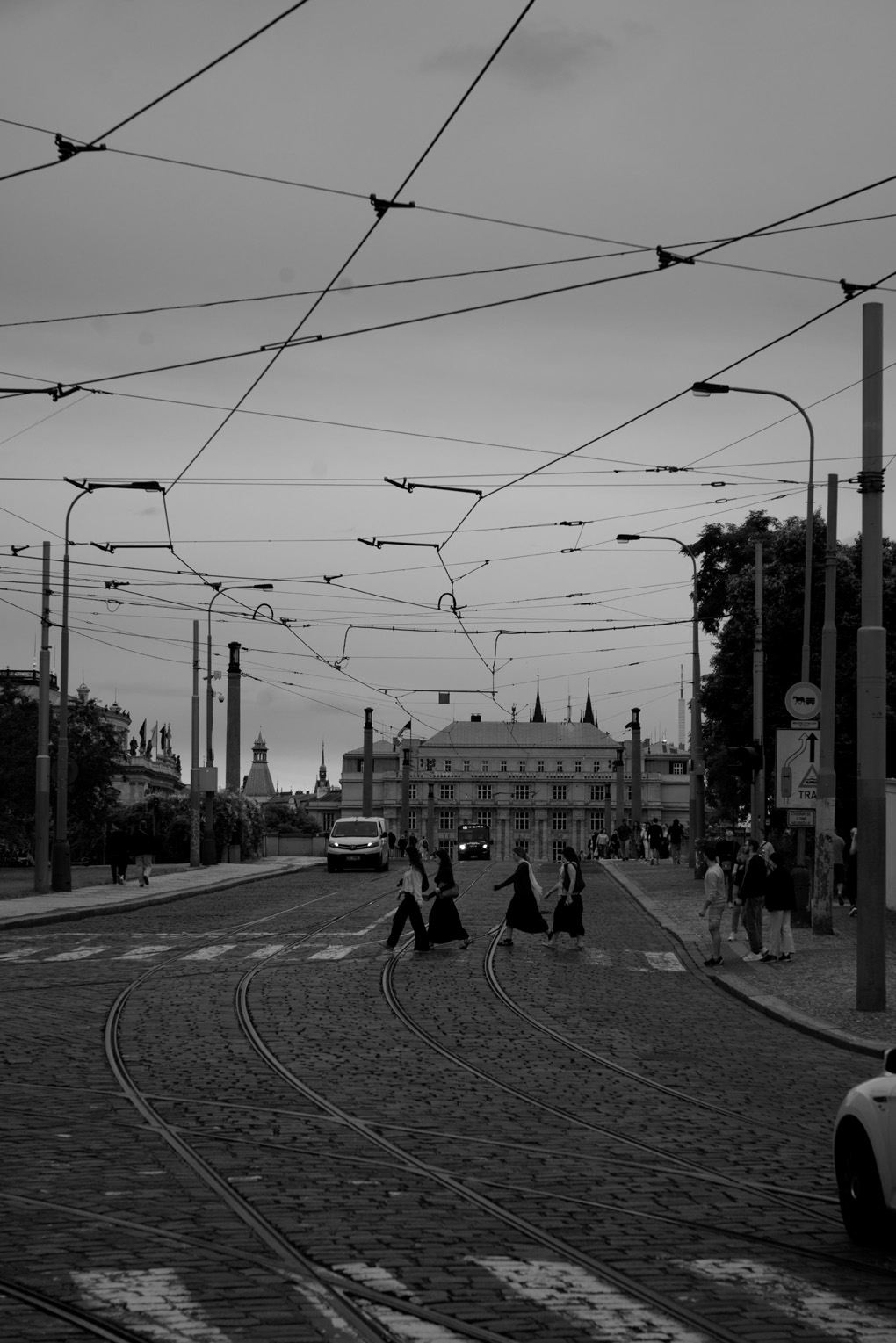 A black and white photo of a family or group of tourists crossing a stone cobbled road on a striped crosswalk. The sky is cut across in all directions by the overhead powerlines for Prague's tram system.