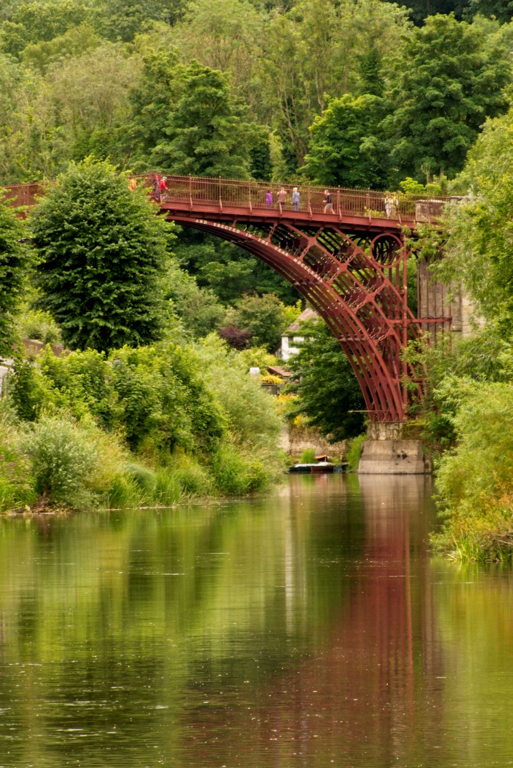 Half of the rusty looking bridge set against verdant green of the trees and faintly reflected in the moving waters of the River Severn