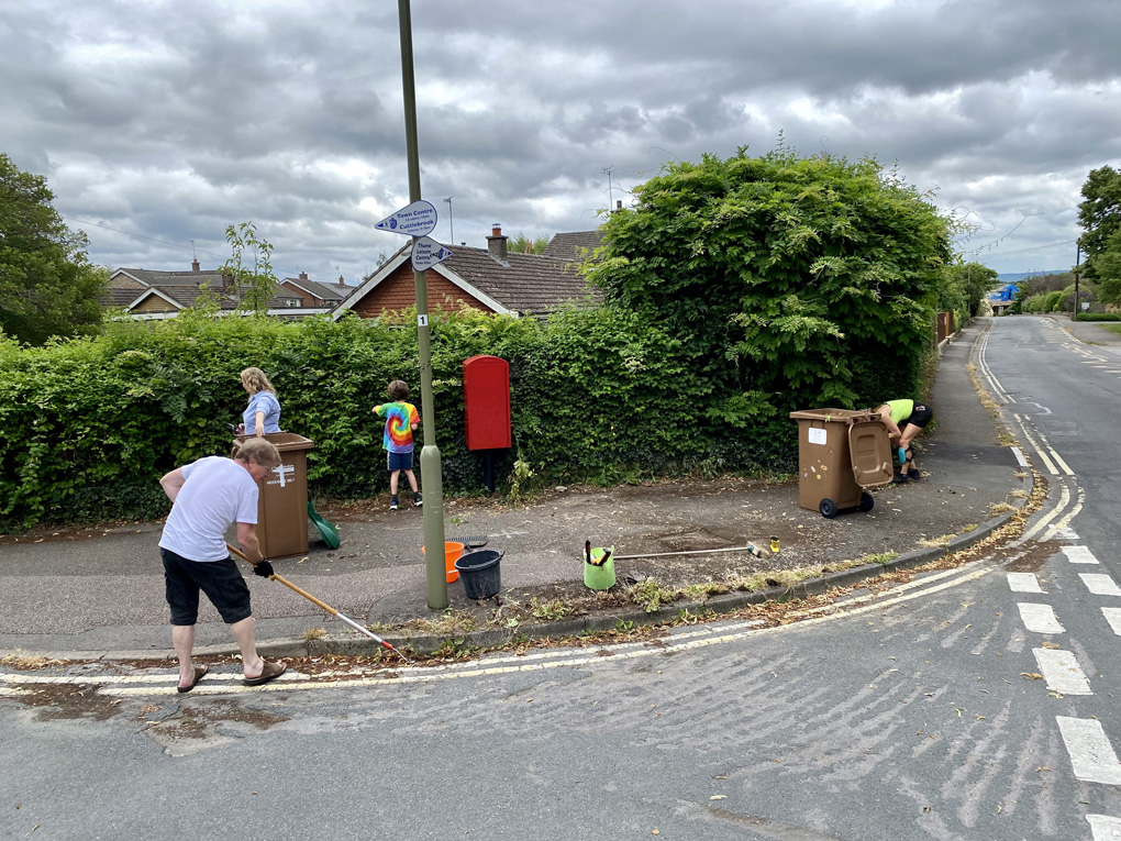 Three adults and a child trimming an overgrown hedge and clearing weeds from a kerb.