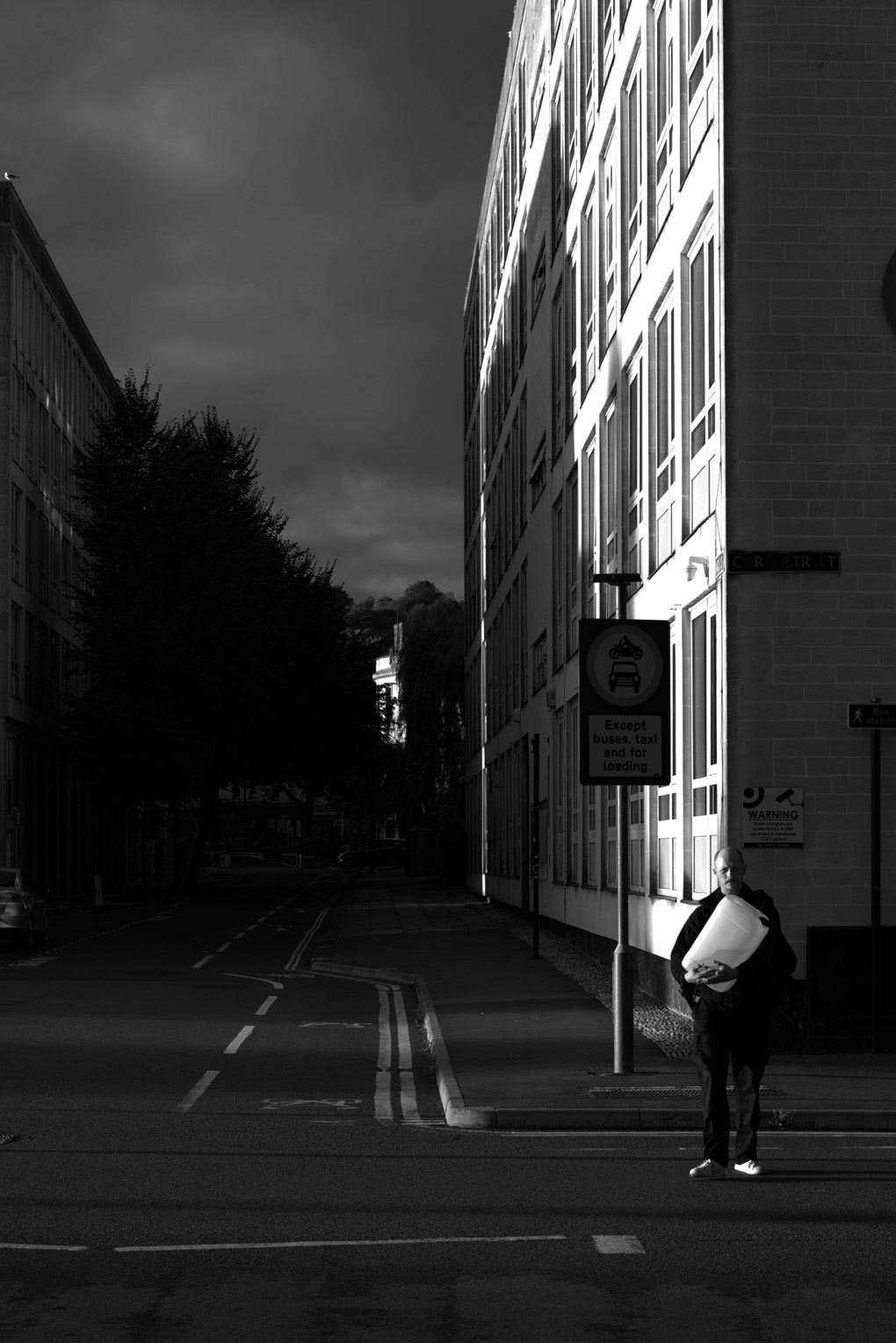 A man in sharp side light, such that half his face is brightly illuminated and the other side is dark. He is standing in front of the corner of a building, similarly contrastingly lit, and the terminator line of the dark and light sides of the building ascends directly from the top of his bald head towards the sky.