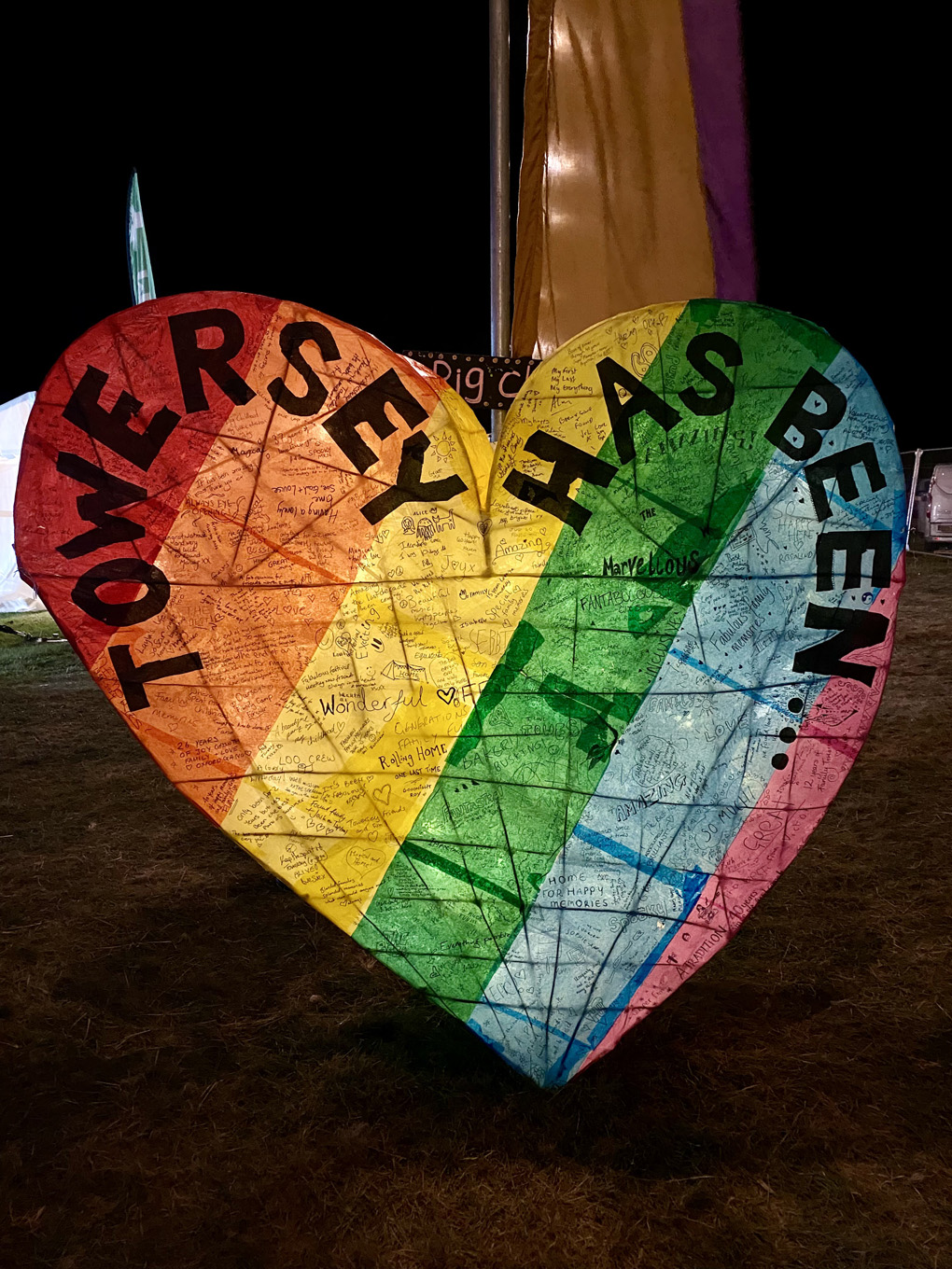 A big heart made of wicker and covered with rainbow tissue paper and lit from within, with black words on saying 