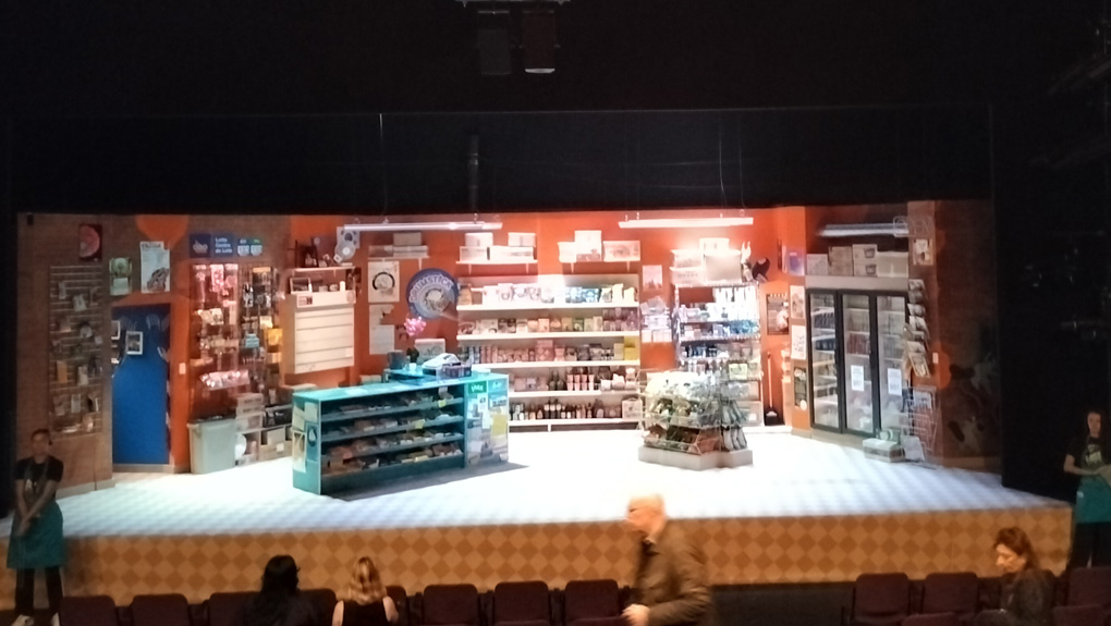 A theatre stage with a set depicting a convnience store. In the foreground we see theatre seats with some people sitting down - above we can see lights. In the middle is the stage itself, with a counter on the left and various well stocked shelves and fridges.