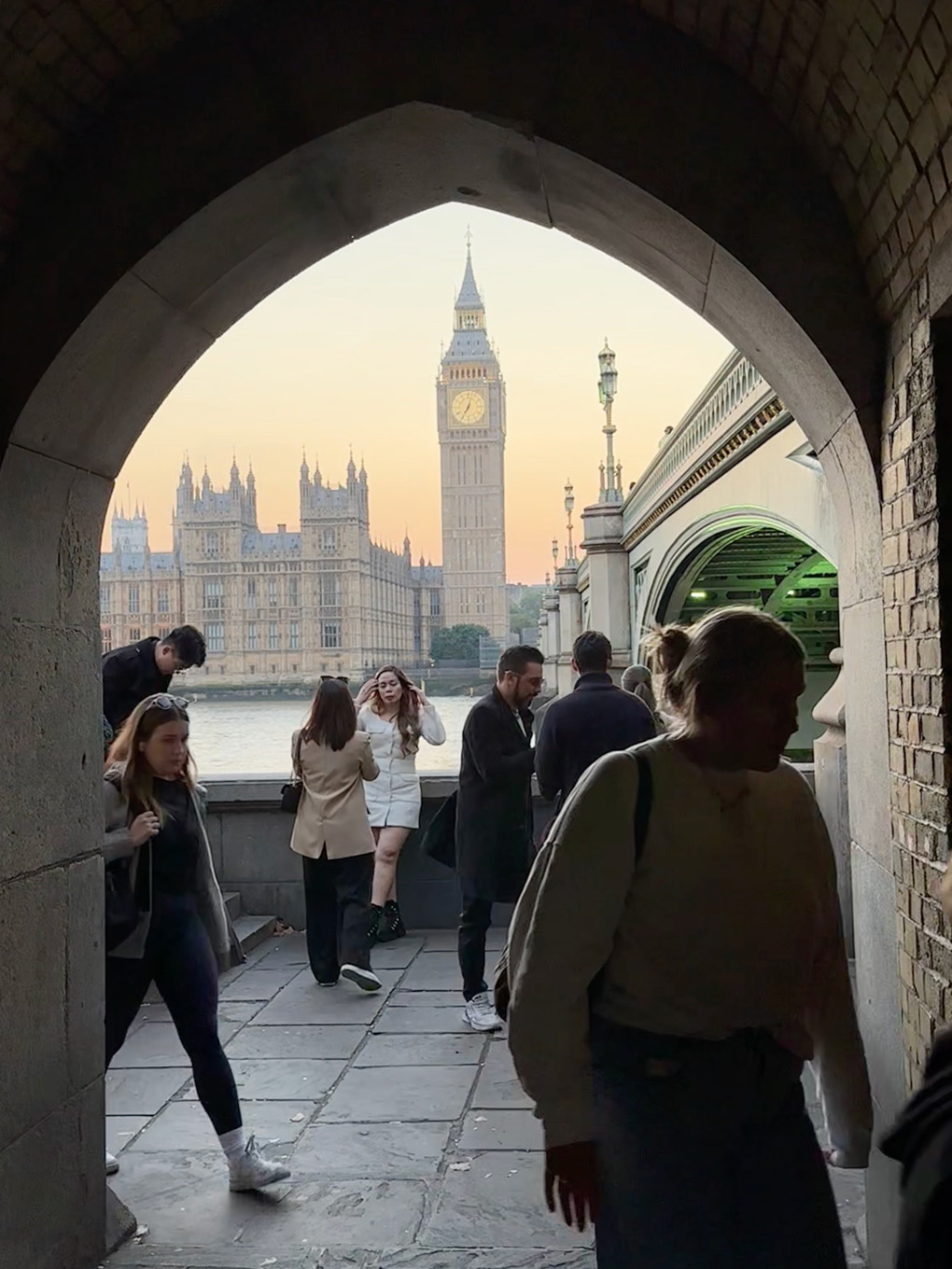 Big Ben and Westminster Bridge at sunset through an arch with people passing by.