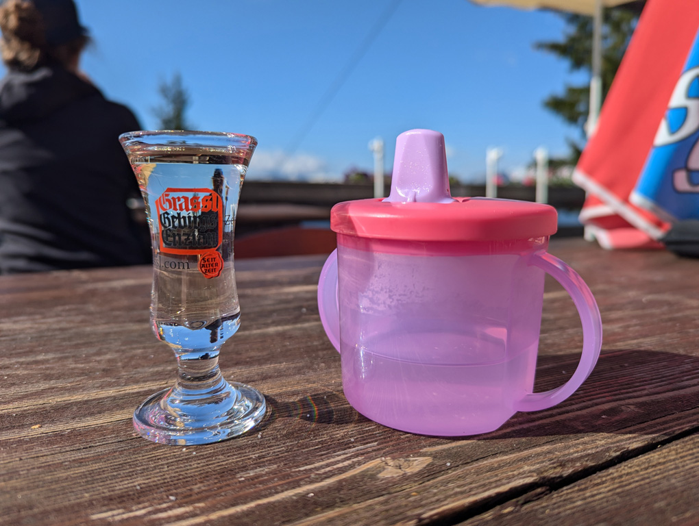 A baby's beaker on a table next to a clear small glass of alcohol