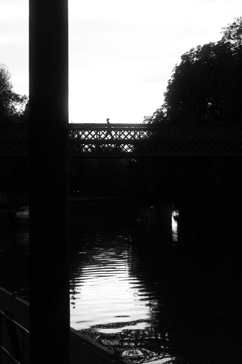 Black and white photo of someone crossing a river over a bridge. Strong contrast makes the figure and bridge all appear in sharp contrast, nearly fully silhouetted.