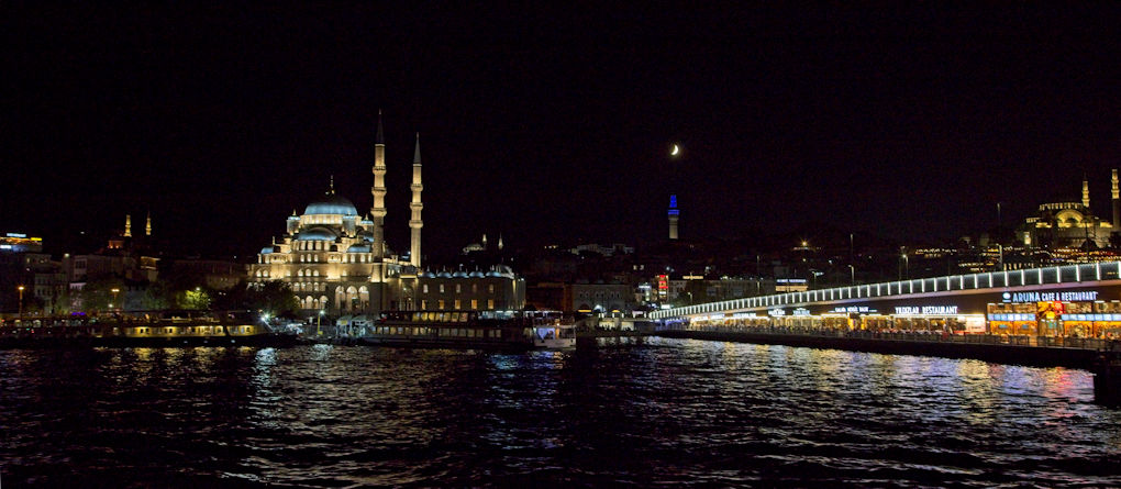 A crescent moon hangs above the nightscape of the European side of Istanbul. The Blue Mosque sits in the centre of the image with the Galata Bridge lining the right hand side. Both have scattered reflections in the choppy water of the Bosphorus., in the foreground.