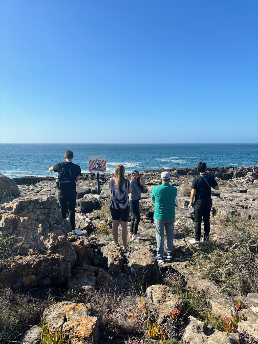 A number of people standing on rocks looking out to sea. Next to them is a sign advising people not to stand there.