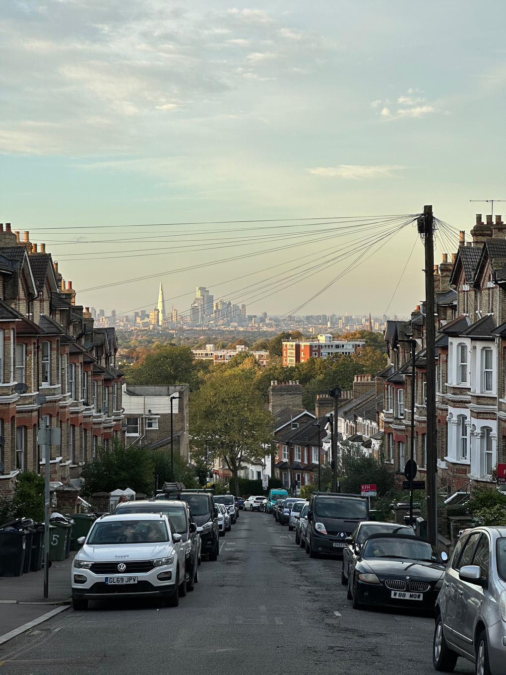 Looking downhill at Central London skyline in distance. Cars parked either side of street. Gherkin, Shard and other buildings visible in far distance.