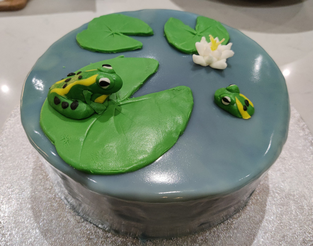 A glossy blue green iced cake with fondant lily pads a lotus flower and two spotty frogs one sitting on a pad the other with its head poking out of the water