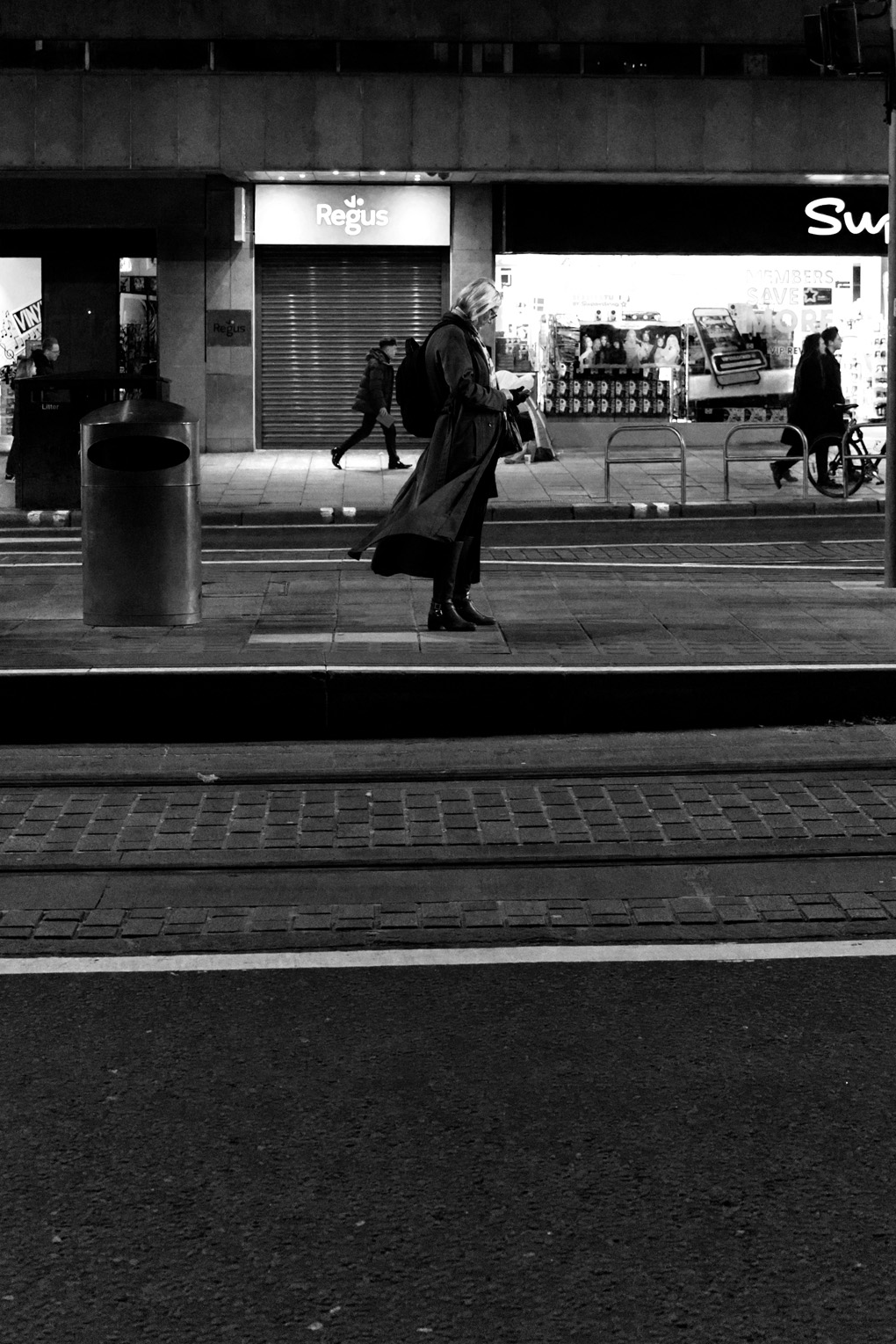 A woman waiting for the tram in Edinburgh on Princes Street. Partially silhouetted, she is in the middleground of the photo on the central island. Wind is blowing her coat a little behind her, while she looks at her phone.