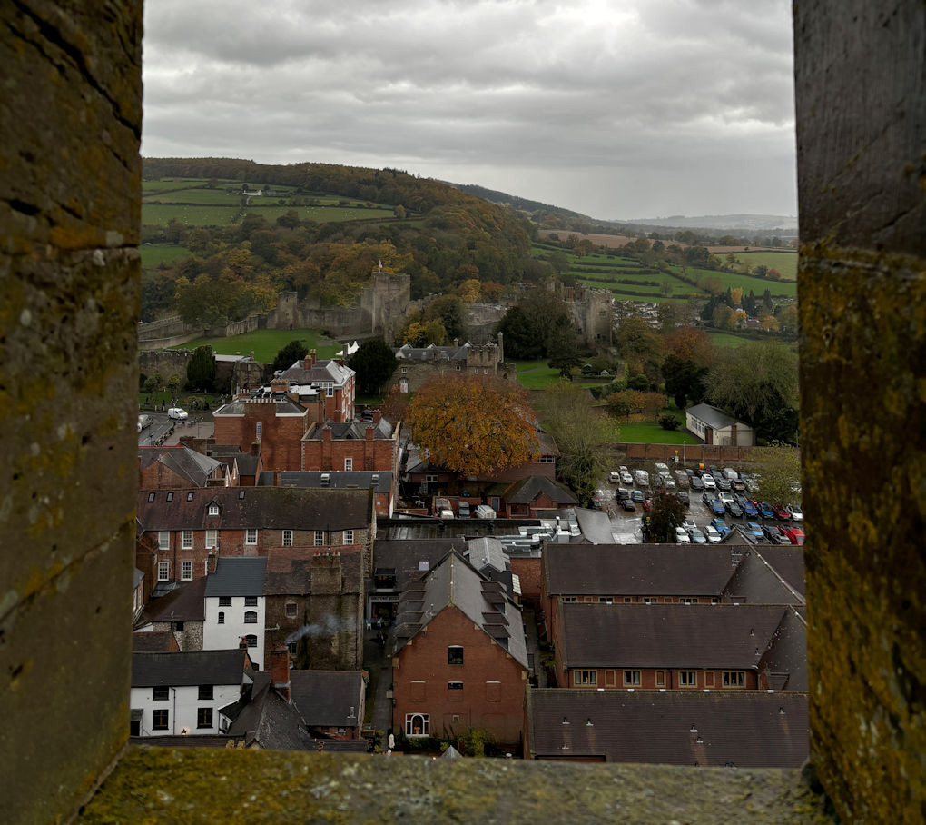 Framed by the castellations of the church tower, the walls and tower of Ludlow castle sit in the middle distance. The tree covered hills beyond merge into the grey, rain filled sky.