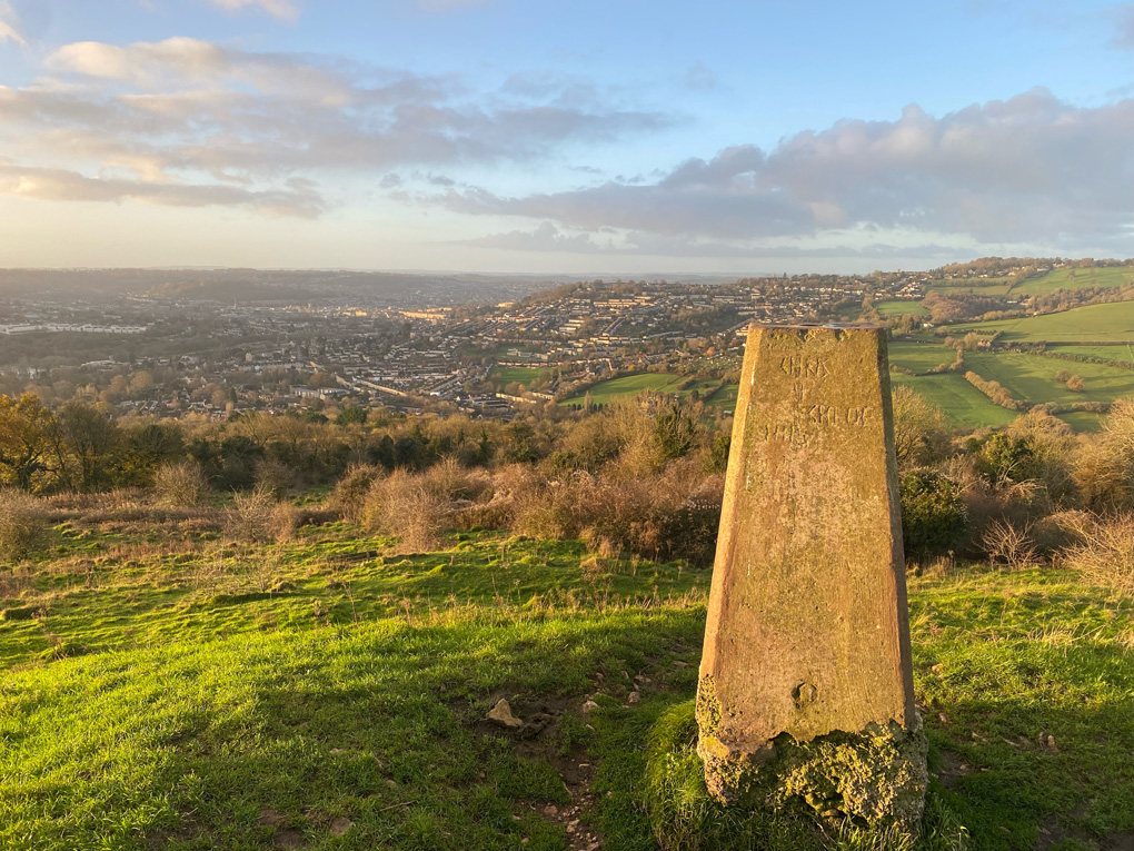 View from the top of Solsbury Hill, lit with low sun. In the foreground is a large stone marker. The city of Bath is visible in the background.