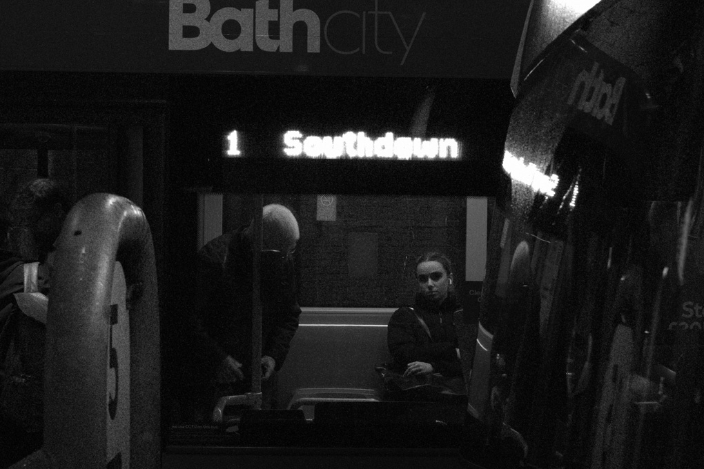 a black and white photo through the side window of a bus waiting at the Bath bus station. A woman seated on the bus looks directly at you while an elderley man is sitting himself down beside her. The glow of the bus's route sign is overexposed and hazy and reflected in the dark windshielf of a second bus parked next to it, off to the side and a little nearer to the viewer.