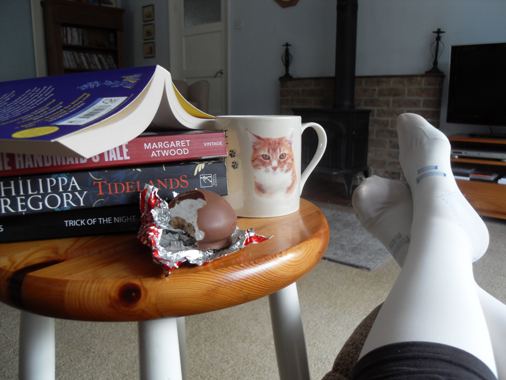 A pair of feet in post-surgery socks with side table heaped with books, tea-mug and choc biscuit.