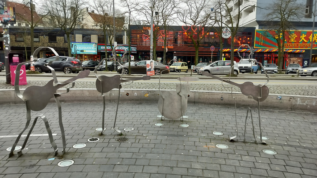 A plaza which faces a busy road and with a street of shops/bars in the background (featuring a Hooters. It's that kind of area). In the foreground are four human-size cookie-cutter-style statues of The Beatles holding guitars or, in the case of Pete Best/Ringo Star, sitting behind a drum kit.