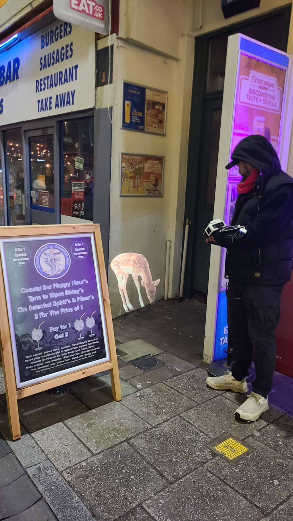 A person dressed in black with a small white projector in their forearm, projecting a deer onto the external, white wall of a takeaway shop.