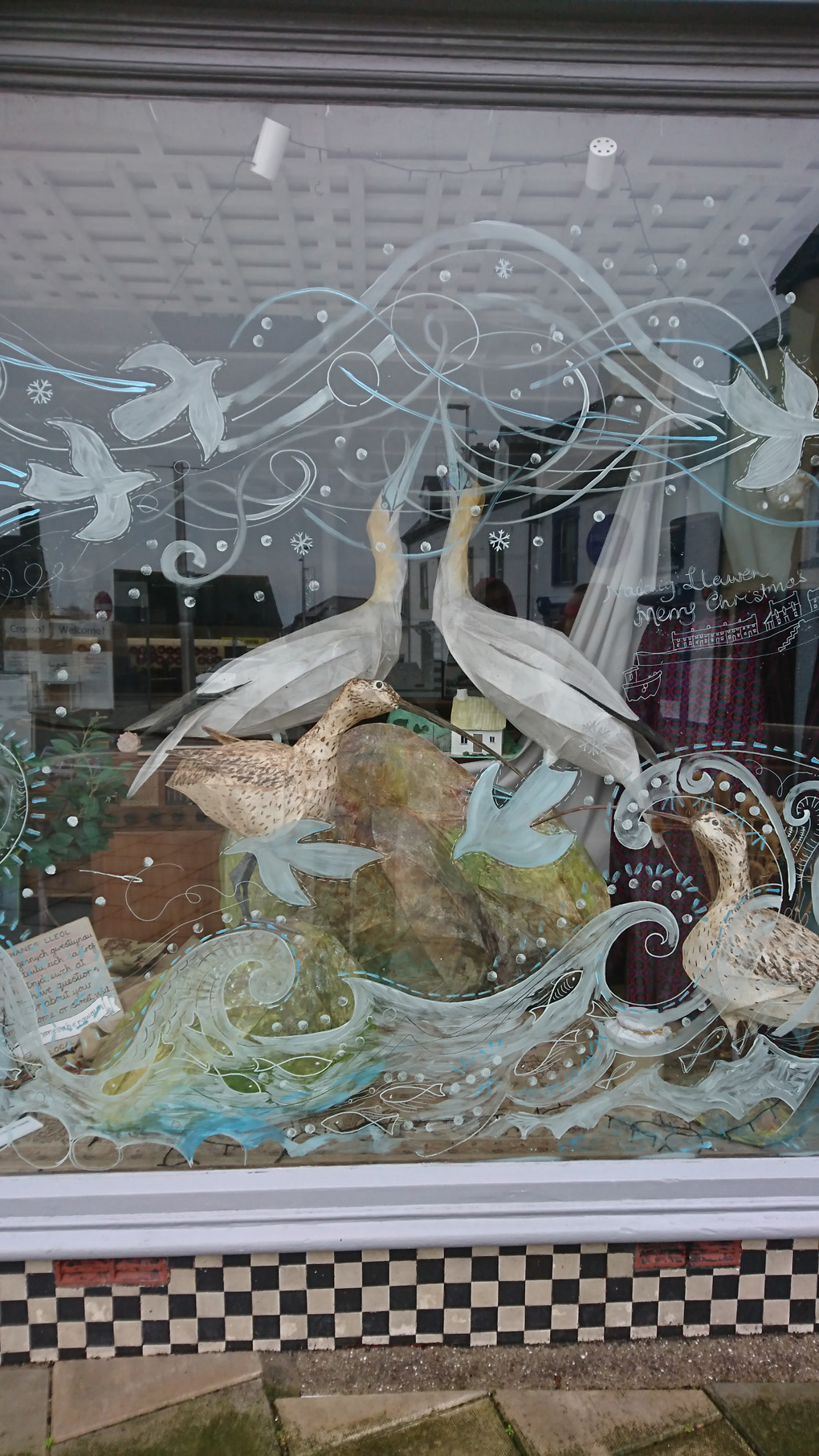 A Christmas window in the coastal town of Fishguard depicting two large gulls and two curlews on a rock rising from the waves with smaller birds flying through swirls of white and dots of sea foam splashed around making for a wintery and cool scene.