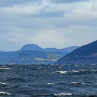 A wave crashes on the stones at the head of loch ness, looking down the loch, into the wind, hills rising beside the water and into the far distance under a brooding sky