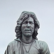 A statue of the three Bee Gees, walking in a line, on the promenade in Douglas. They are walking away from the sea, which is visible in the background.