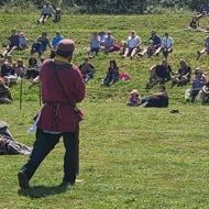 A grass amphitheater with a rope cordon, blue sky above. Spectators sit watching a Viking battle, many lay 