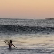 Large waves rolling in towards a beach at sunset. Three people are wading into the water towards the waves, arms outstretched