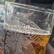 Two people holding half-drunk glasses of beer. On the background, our of focus, is a baby peering over the top of a backpack carrier