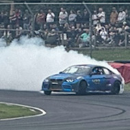 Two sports cars - purple in the foreground, blue in the background - drifting around a wide curve of a race track in rural England