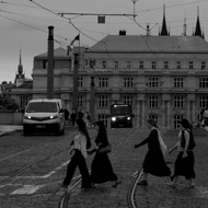 A black and white photo of a family or group of tourists crossing a stone cobbled road on a striped crosswalk. The sky is cut across in all directions by the overhead powerlines for Prague's tram system.