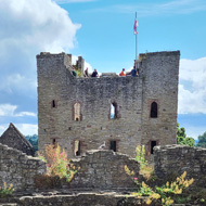 The view from a walkway part way up the walls, looking through Ludlow Castle, towards the Keep with a sunny blue sky and fluffy clouds There is a wooden railing in the fireground and lots of green vegetetaion grwoing out of the Castle's stone walls..