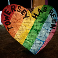 A big heart made of wicker and covered with rainbow tissue paper and lit from within, with black words on saying 