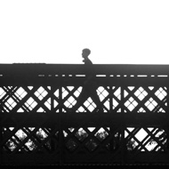 Black and white photo of someone crossing a river over a bridge. Strong contrast makes the figure and bridge all appear in sharp contrast, nearly fully silhouetted.