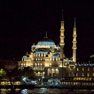 A crescent moon hangs above the nightscape of the European side of Istanbul. The Blue Mosque sits in the centre of the image with the Galata Bridge lining the right hand side. Both have scattered reflections in the choppy water of the Bosphorus., in the foreground.