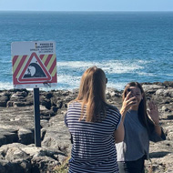 A number of people standing on rocks looking out to sea. Next to them is a sign advising people not to stand there.