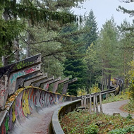 An autumnal and wet day - a concrete bobsleigh track lies ahead, covered in pine needles. There are bends which show how the track curves upwards as it goes around corners. At the bends the track is covered in graffiti.