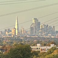 Looking downhill at Central London skyline in distance. Cars parked either side of street. Gherkin, Shard and other buildings visible in far distance.