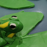 A glossy blue green iced cake with fondant lily pads a lotus flower and two spotty frogs one sitting on a pad the other with its head poking out of the water