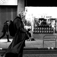 A woman waiting for the tram in Edinburgh on Princes Street. Partially silhouetted, she is in the middleground of the photo on the central island. Wind is blowing her coat a little behind her, while she looks at her phone.