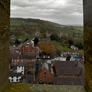 Framed by the castellations of the church tower, the walls and tower of Ludlow castle sit in the middle distance. The tree covered hills beyond merge into the grey, rain filled sky.